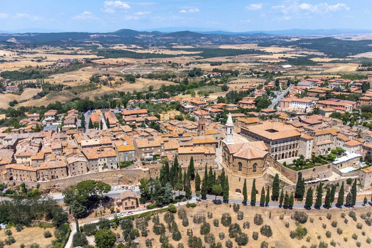 Stazione ferroviaria tipica della Toscana, treno regionale in sosta, viaggiatori con valigie, atmosfera rilassata e paesaggio collinare sullo sfondo